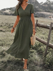 Abito lungo verde oliva estivo da donna, con scollo a V e maniche corte, indossato in un ambiente naturale. Accessoriato con borsa di paglia.