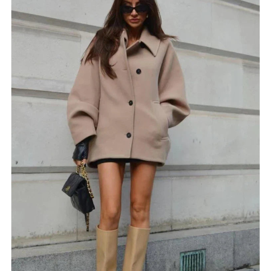 Donna con cappotto beige oversize, stivali alti beige, borsa nera con catena dorata, occhiali da sole, posa su sfondo urbano. Moda autunnale elegante.