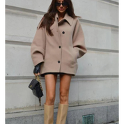 Donna con cappotto beige oversize, stivali alti beige, borsa nera con catena dorata, occhiali da sole, posa su sfondo urbano. Moda autunnale elegante.