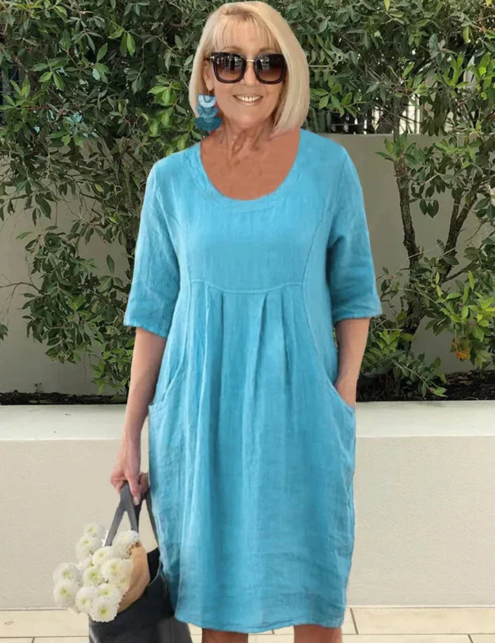 Donna con abito estivo blu in lino, occhiali da sole, orecchini turchesi, borsa nera e fiori bianchi, sfondo di piante verdi.