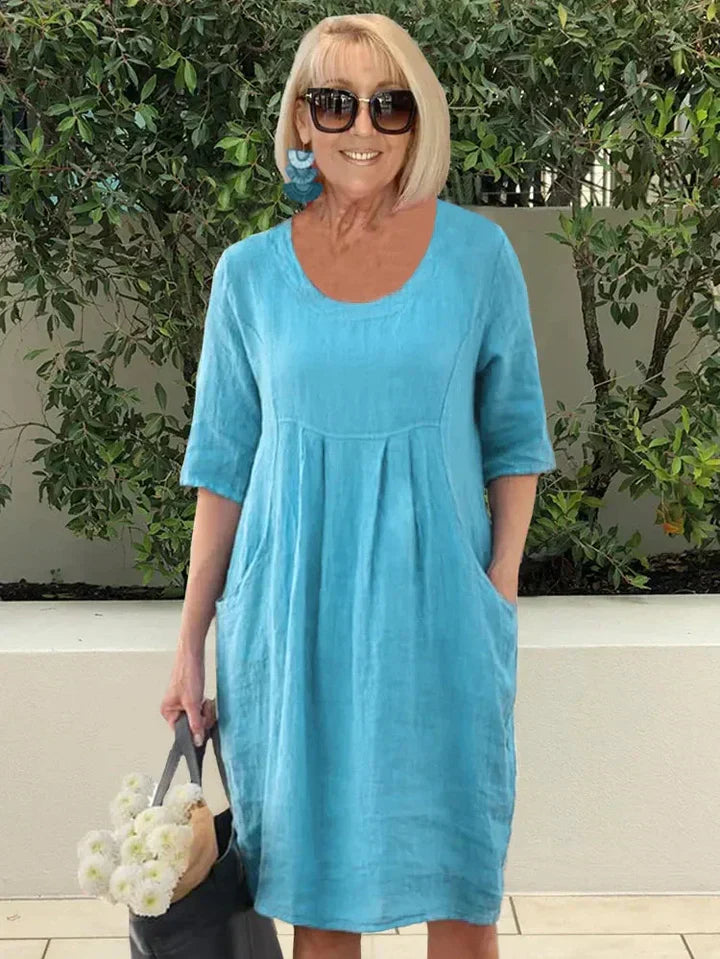 Donna con abito estivo blu in lino, occhiali da sole, orecchini turchesi, borsa nera e fiori bianchi, sfondo di piante verdi.