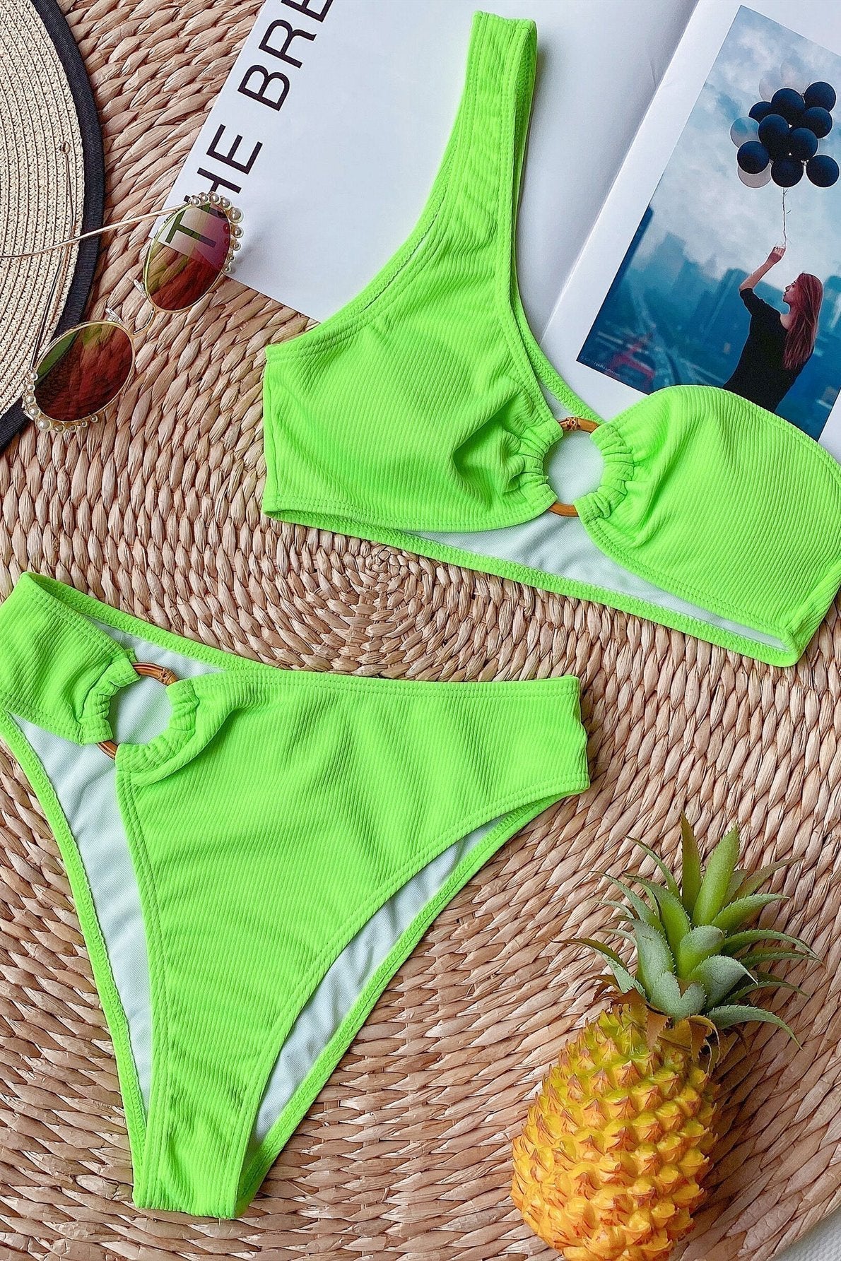 Bikini verde fluo a costine con anello decorativo su sfondo estivo, ananas, occhiali da sole e rivista. Moda mare, costumi da bagno trendy.