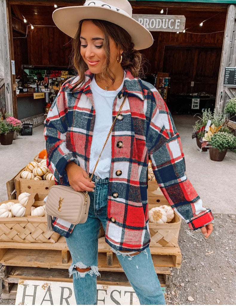 Donna con camicia a quadri rossi e blu, jeans strappati, borsa beige, cappello beige, davanti a un mercato agricolo con zucche e fiori.