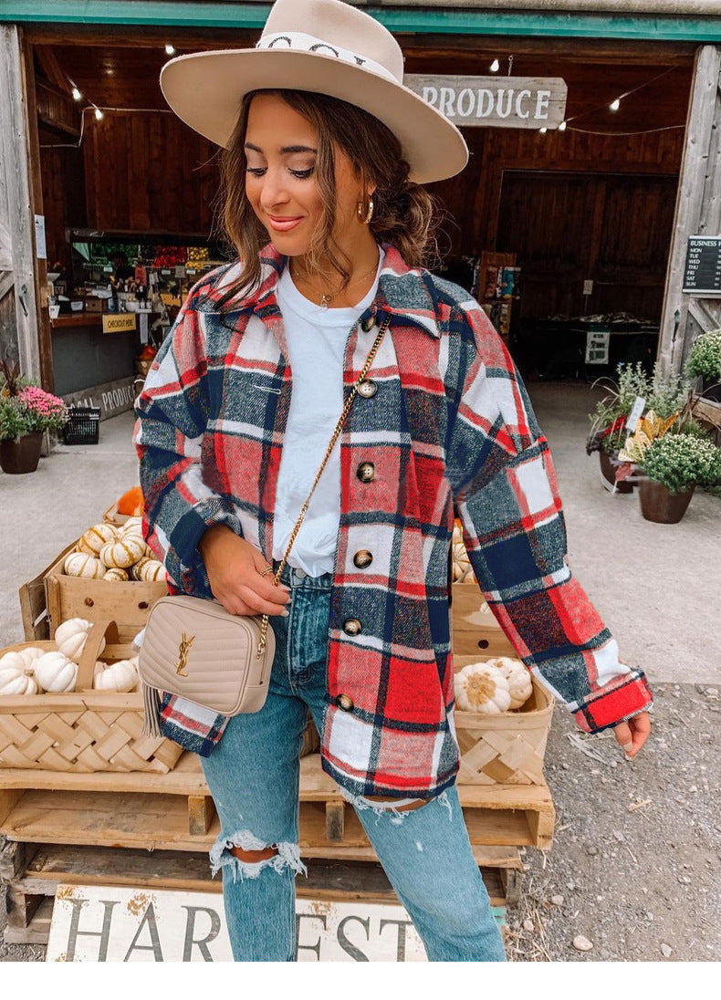 Donna con camicia a quadri rossi e blu, jeans strappati, borsa beige, cappello beige, davanti a un mercato agricolo con zucche e fiori.