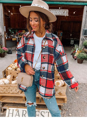 Donna con camicia a quadri rossi e blu, jeans strappati, borsa beige, cappello beige, davanti a un mercato agricolo con zucche e fiori.