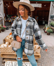 Donna con cappotto a quadri grigio, jeans strappati e borsa beige, davanti a un mercato agricolo con zucche e fiori. Moda autunnale casual.