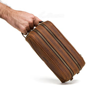 Mano che tiene un elegante beauty case in pelle marrone con doppia cerniera, ideale per viaggi e organizzazione accessori. Design compatto e resistente.