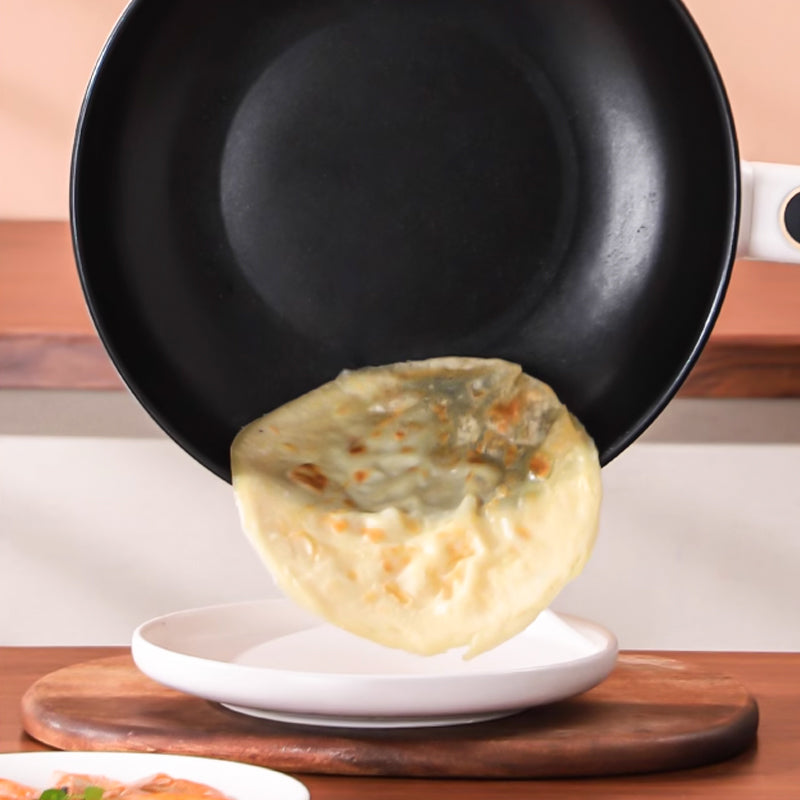 Padella antiaderente nera con manico bianco mentre versa una crepe dorata su un piatto bianco, cucina moderna, utensili da cucina essenziali.