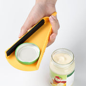 Utensile apribarattoli giallo in plastica, impugnato da una mano, utilizzato per aprire un barattolo di vetro con coperchio verde. Accessorio cucina pratico.