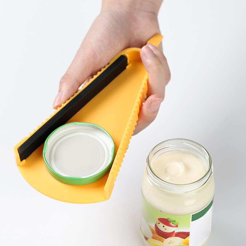 Utensile apribarattoli giallo in plastica, impugnato da una mano, utilizzato per aprire un barattolo di vetro con coperchio verde. Accessorio cucina pratico.