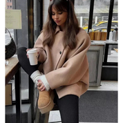 Donna con cappotto beige oversize, leggings neri e stivali UGG, seduta in un caffè con caffè da asporto. Look casual invernale alla moda.