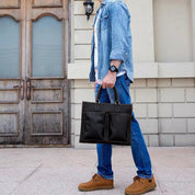 Uomo con borsa a mano in pelle nera, indossa jeans e camicia di jeans, scarpe marroni. Moda casual maschile, accessori eleganti.