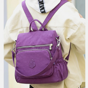 Zaino viola in tessuto resistente, con tasche multiple e cerniere, indossato su giacca beige. Ideale per scuola, viaggio, tempo libero.