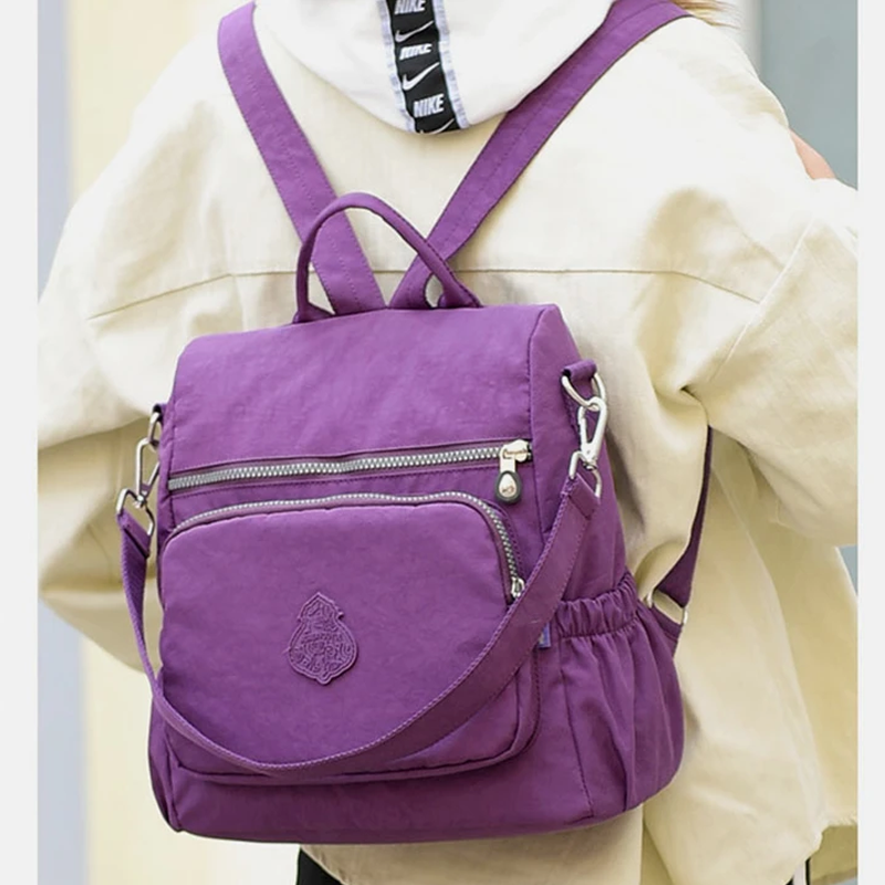 Zaino viola in tessuto resistente, con tasche multiple e cerniere, indossato su giacca beige. Ideale per scuola, viaggio, tempo libero.