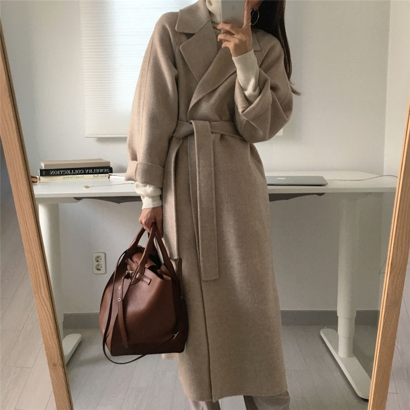 Donna con cappotto beige lungo e cintura, maglione bianco, borsa marrone, in piedi davanti a uno specchio in una stanza moderna e minimalista.