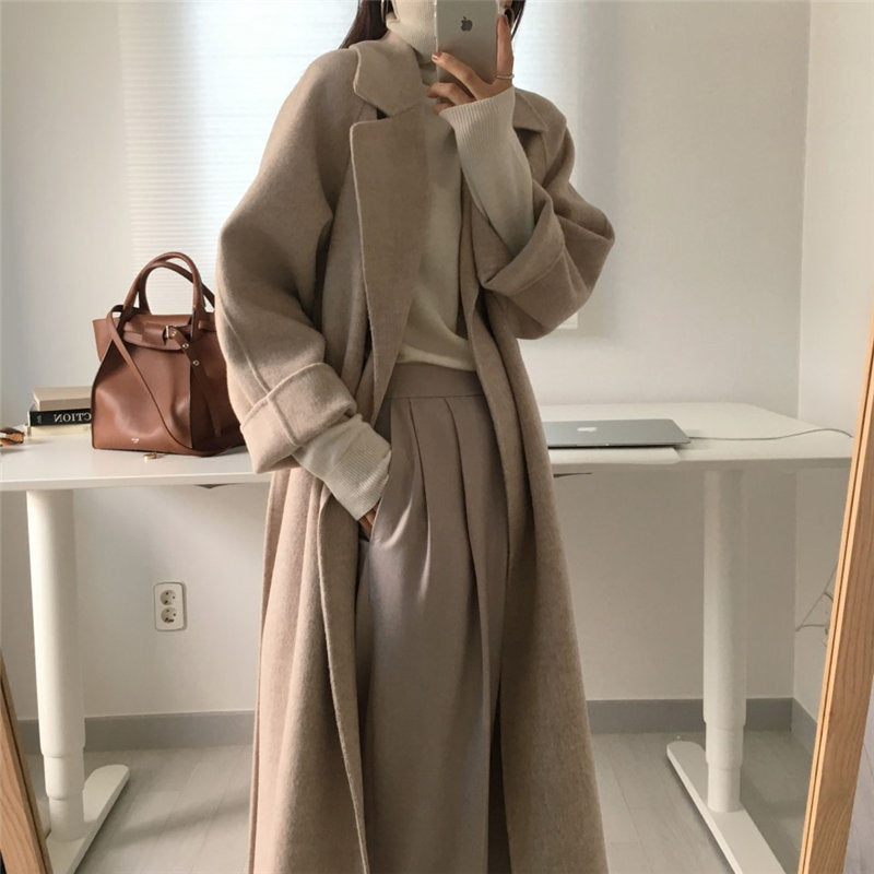 Cappotto lungo beige elegante, abbinato a maglione e pantaloni larghi, davanti a scrivania moderna con borsa in pelle marrone. Moda autunnale donna.
