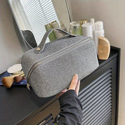 Borsa da toilette grigia con cerniera, tessuto intrecciato, su mobile bagno moderno. Accessori da bagno sullo sfondo. Elegante e funzionale.