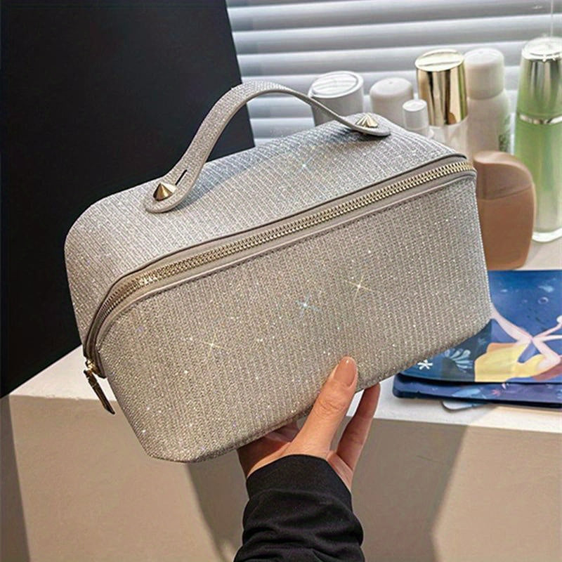 Borsa cosmetica grigia scintillante con cerniera, manico superiore e dettagli metallici, su sfondo di prodotti per la cura della pelle.