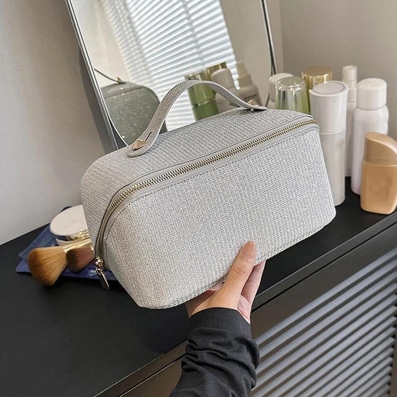 Borsa cosmetica grigia con cerniera, manico superiore, su mobile da bagno. Accessori trucco e specchio sullo sfondo. Elegante e funzionale.