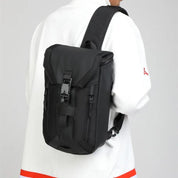 Zaino nero a tracolla unisex, design moderno e compatto, con chiusura a fibbia e tasche con zip, indossato su giacca bianca. Accessorio moda urbano.