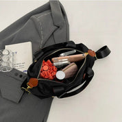 Borsa nera aperta su giacca grigia, contenente smartphone, portafoglio, rossetto, occhiali e libro. Accessori moda donna eleganti e pratici.