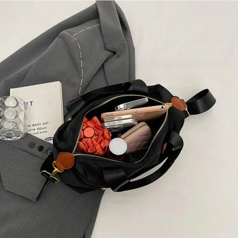 Borsa nera aperta su giacca grigia, contenente smartphone, portafoglio, rossetto, occhiali e libro. Accessori moda donna eleganti e pratici.