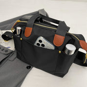 Borsa a tracolla nera in nylon con dettagli in pelle marrone, tasche esterne contenenti smartphone e cosmetici, su sfondo di abbigliamento grigio.