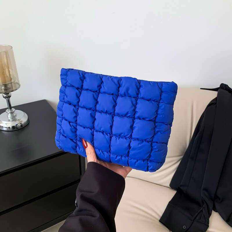 Mano che tiene una borsa trapuntata blu su un divano beige, accanto a un tavolino nero con una candela. Accessorio moda elegante e moderno.