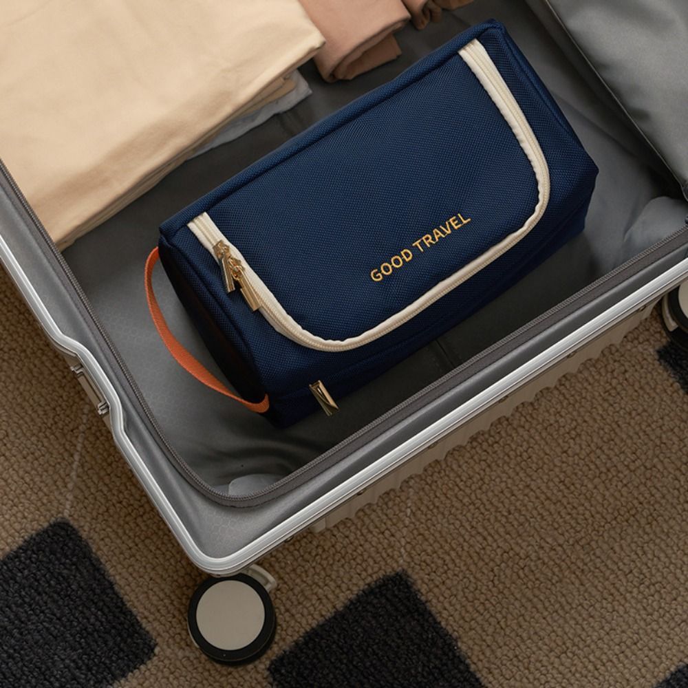 Valigia aperta con beauty case blu "Good Travel", cerniera dorata, su tappeto beige. Accessori viaggio, organizzazione bagaglio, praticità.
