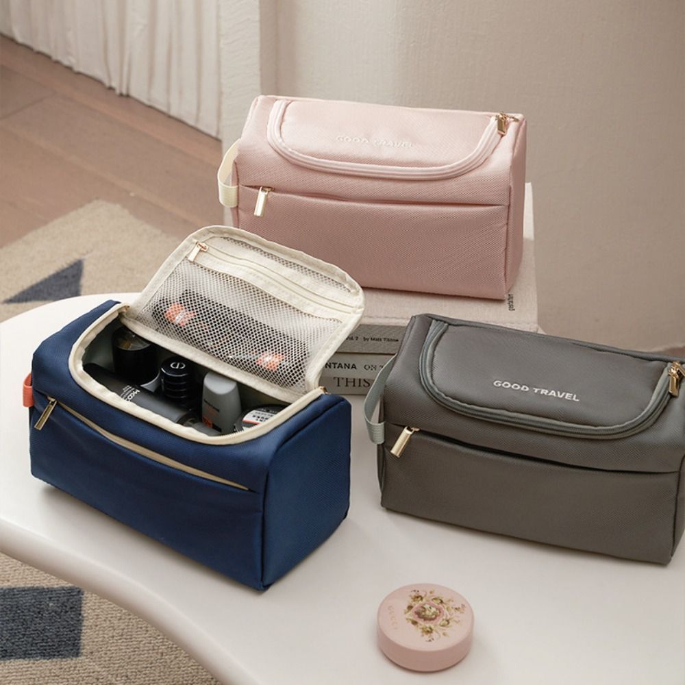 Tre beauty case da viaggio in tessuto, colori rosa, blu e grigio, con cerniere dorate e scomparti interni, su tavolo in legno chiaro.