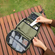 Borsa da viaggio verde aperta su panchina di legno, con scomparti contenenti flaconi e accessori per la cura personale. Mani che organizzano gli oggetti.