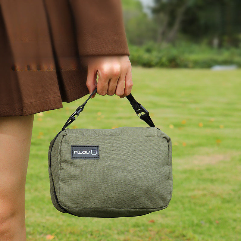 Borsa a tracolla verde militare, compatta e resistente, tenuta in mano su sfondo erboso. Ideale per viaggi, escursioni e uso quotidiano.