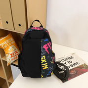 Zaino nero con scritte colorate su tavolo, accanto a sacchetto di popcorn e giornale. Design moderno, ideale per scuola o viaggio.