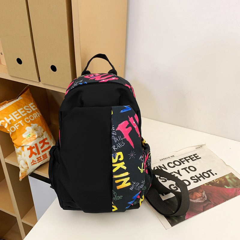 Zaino nero con scritte colorate su tavolo, accanto a sacchetto di popcorn e giornale. Design moderno, ideale per scuola o viaggio.
