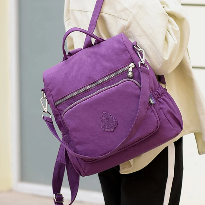 Zaino viola in tessuto resistente, con tasche multiple e cerniere, indossato su una spalla. Ideale per scuola, viaggio, uso quotidiano.