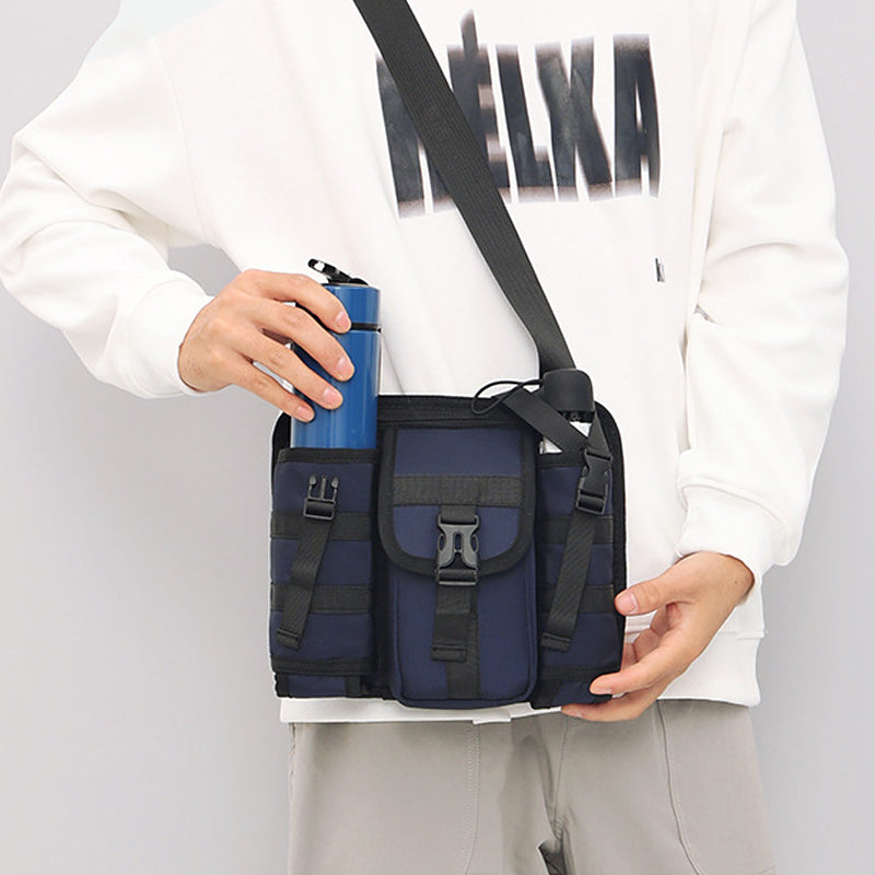 Borsa a tracolla unisex blu scuro con tasche multiple, indossata da una persona in felpa bianca, che tiene una borraccia blu. Accessorio moda casual.