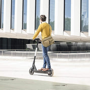 Uomo su monopattino elettrico nero, indossa maglione giallo e jeans, davanti a edificio moderno. Mobilità urbana sostenibile.