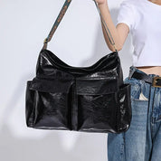 Borsa a tracolla nera in pelle lucida con tasche frontali, indossata da una donna in jeans e maglietta bianca. Accessorio moda elegante e spazioso.