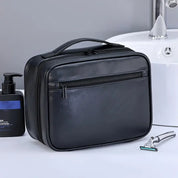 Borsa da toilette in pelle nera con cerniera, manico superiore, accanto a rasoio e dispenser sapone. Elegante e pratica per viaggi.
