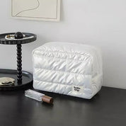 Borsa da viaggio bianca trapuntata su tavolo nero, accanto a vassoio con perle e profumo. Design elegante, ideale per organizzare accessori.