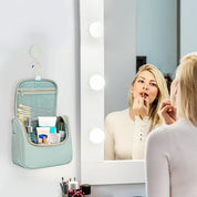 Donna si trucca davanti a specchio illuminato, accanto a borsa da toilette verde acqua appesa, contenente cosmetici e accessori per il trucco.