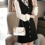 Donna in abito nero elegante con camicia bianca, borsa a tracolla bianca con catena dorata, seduta su poltrona moderna vicino a finestra.
