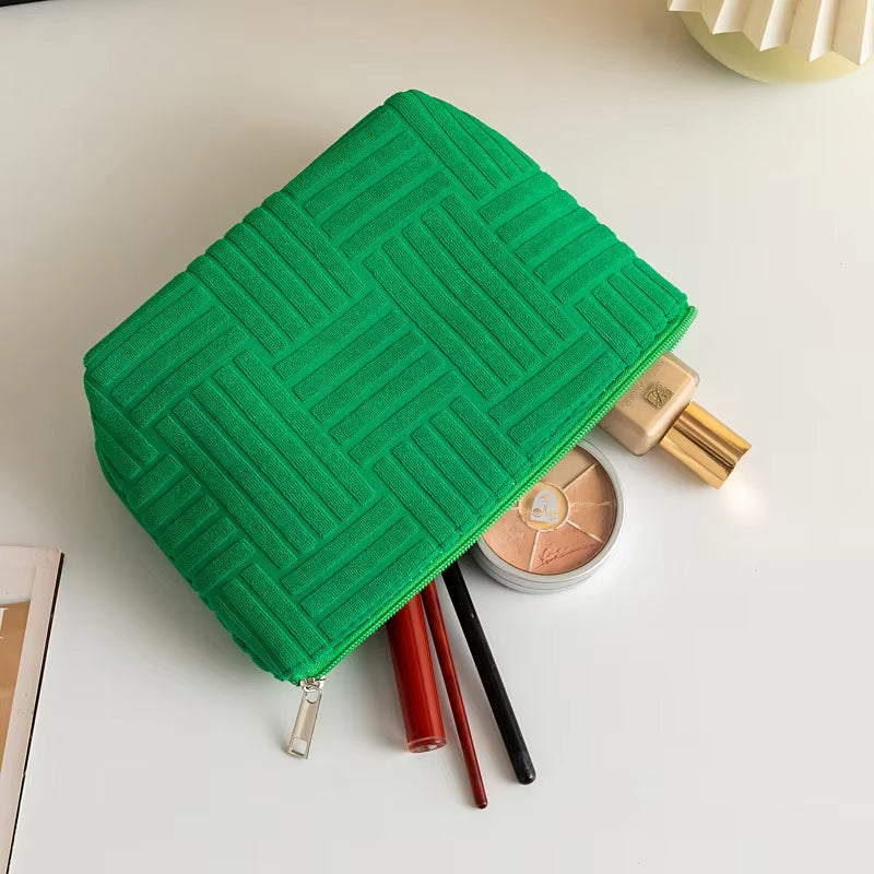 Astuccio verde in tessuto con motivo geometrico, contenente trucchi: fondotinta, palette ombretti, rossetto rosso e matite occhi su tavolo bianco.