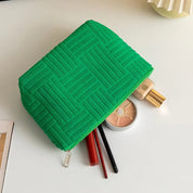 Astuccio verde in tessuto con motivo geometrico, contenente trucchi: fondotinta, palette ombretti, rossetto rosso e matite occhi su tavolo bianco.