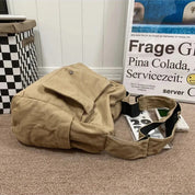 Borsa a tracolla beige in tela su pavimento marrone, accanto a scatola a scacchi e poster. Design casual, ideale per uso quotidiano e viaggi.