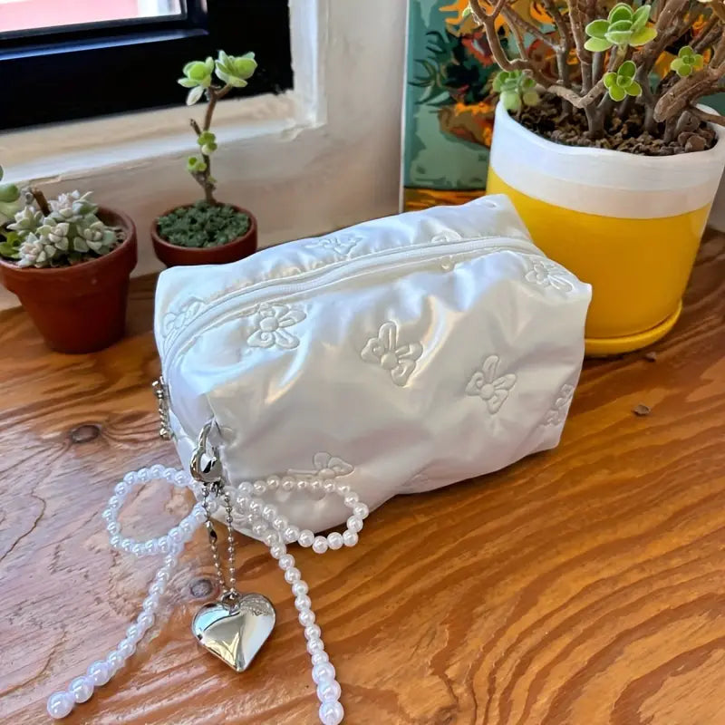 Astuccio bianco in raso con ricami floreali, decorato con perline e ciondolo a cuore, su tavolo di legno accanto a piante grasse in vaso.