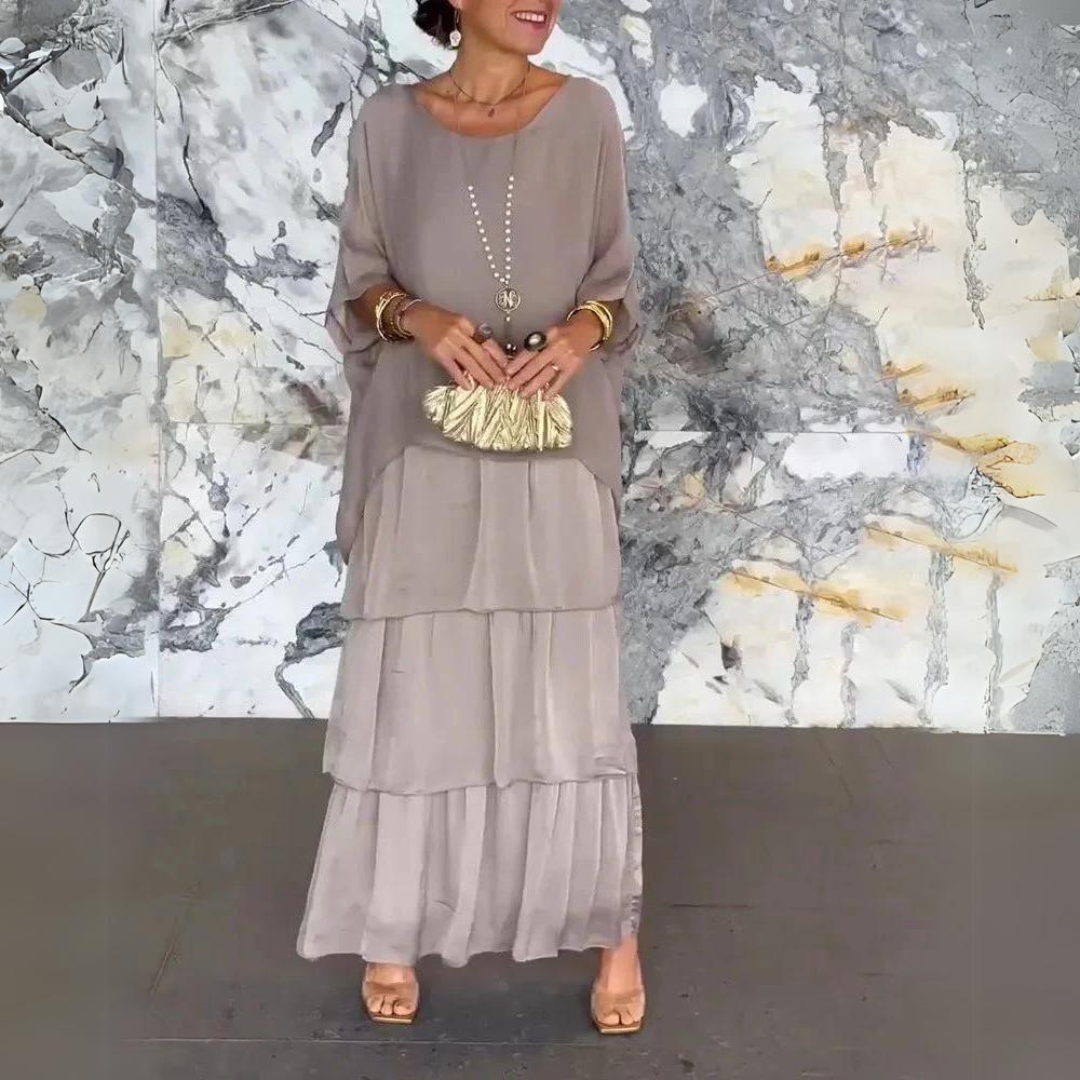 Abito lungo elegante grigio con balze, accessoriato con collana e pochette dorata, indossato da donna su sfondo marmo. Moda femminile sofisticata.