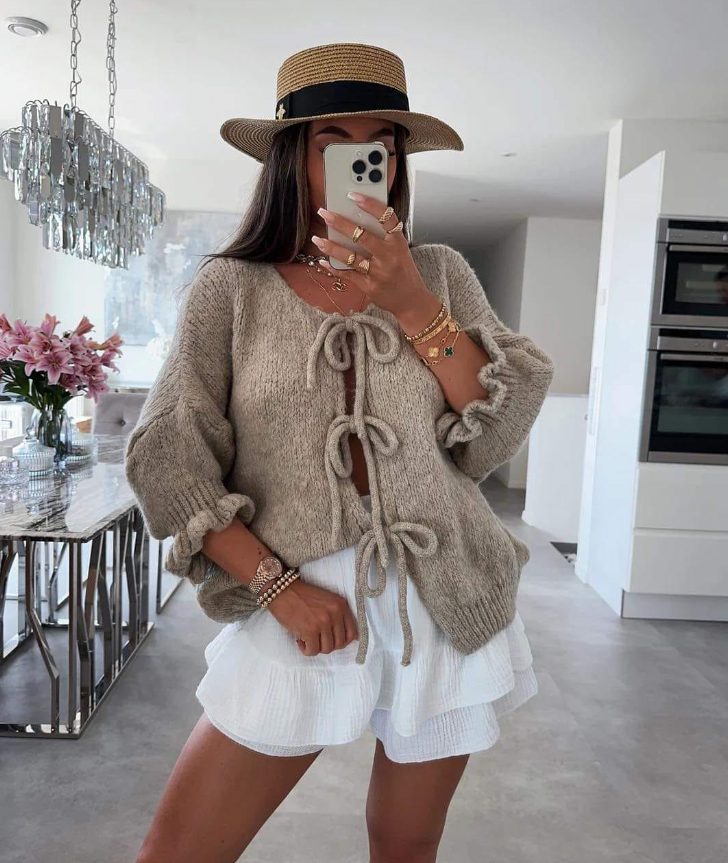 Donna con cappello di paglia, maglione beige con fiocchi, gonna bianca a balze, scatta selfie in cucina moderna. Moda estiva, abbigliamento casual chic.