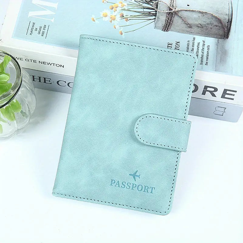 Custodia passaporto in pelle azzurra su tavolo bianco, accanto a libro e pianta. Design elegante, ideale per viaggiatori. Accessorio viaggio essenziale.