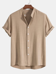 Camicia beige da uomo in lino, maniche corte, colletto alla coreana, bottoni bianchi, stile casual estivo, appesa su gruccia di legno.